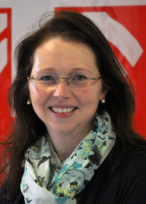 Eine Person mit langen Haaren, Perlenohrringen und einem gemusterten Schal. Der Hintergrund besteht aus einem roten und weißen Design. 